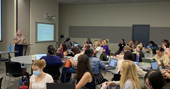 Dr. Amy Ardell is presenting to educators sitting at round tables in a conference room