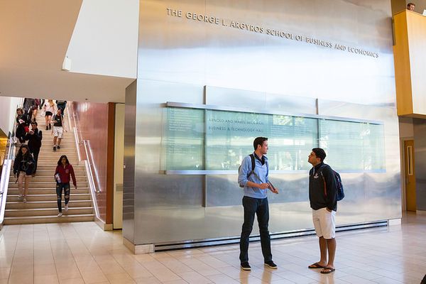 Two students have a discussion near a staircase in a wide, bright hallway.