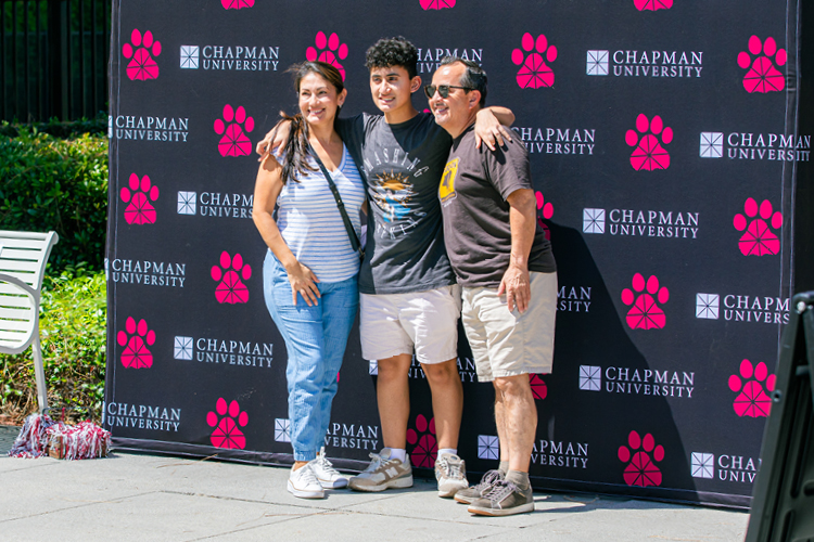 A chapman student stands in front of a Chapman branded banner with both arms around their parents.