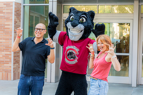 Two Chapman alumni pose with the Pete the Panther mascot with their hands up in the "paws up" stance.