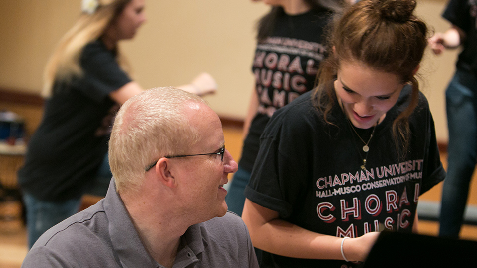 Seated white male with blonde short-cropped hair wearing a gray polo shirt talking to a young white female with brown pony-tail wearing a black choral camp t-shirt.