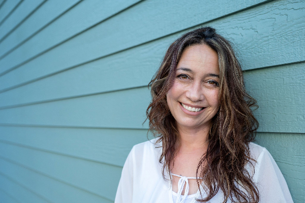 A woman with long brown, wavy hair and wearing a white shirt is smiling while standing in front of a light blue shiplap wall.