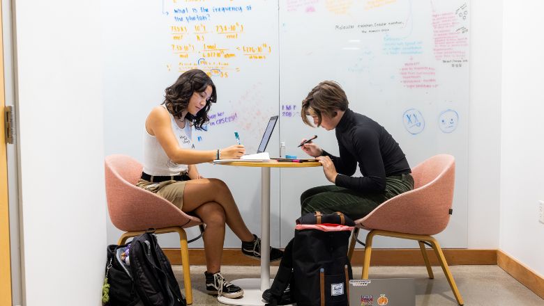 Two students sitting in front of a white board studying. 