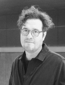 Black and white photo of a white man with glasses and curly hair. 