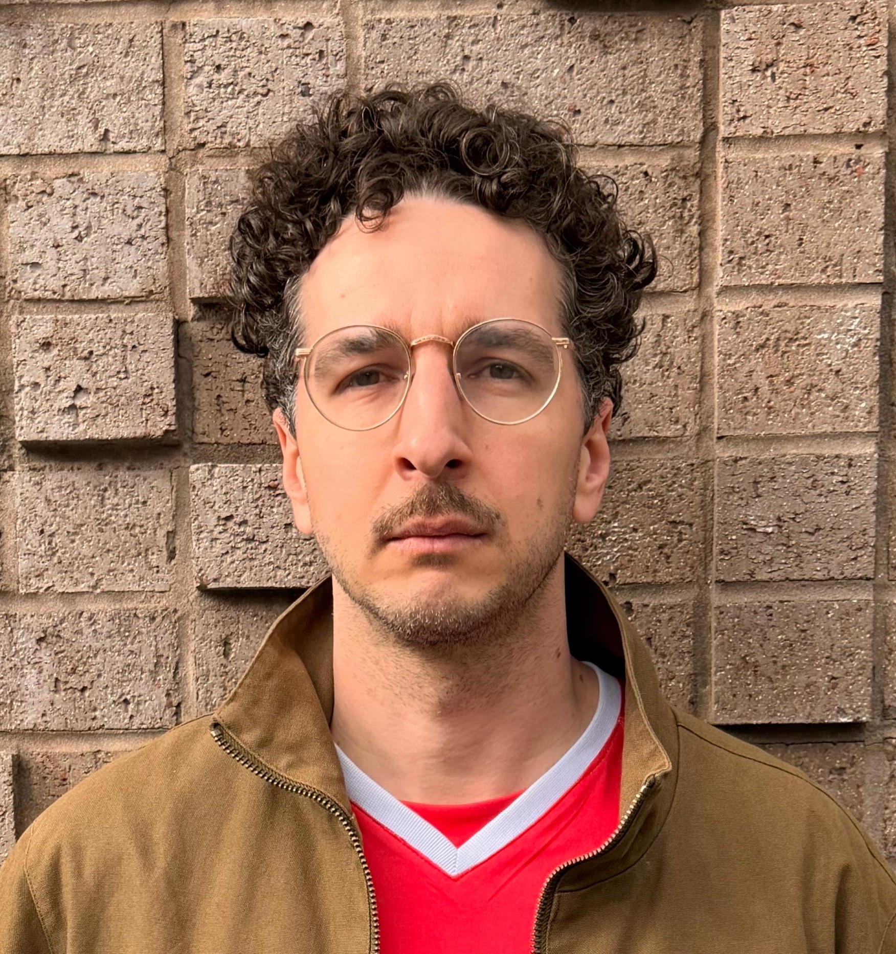 White man with glasses and stash with curly hair standing infront of a wall.