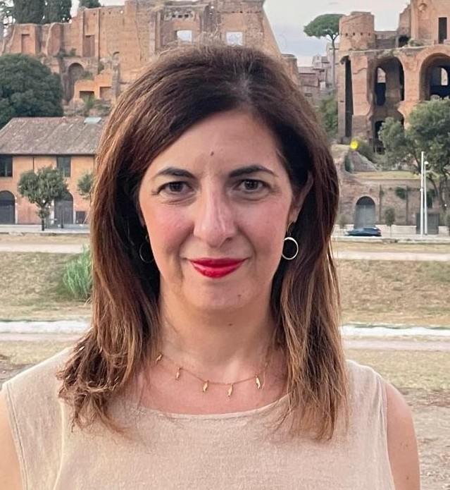 Woman with brown hair and red lips, beigh shirt and necklace.