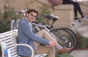 A student wearing formal attire sits on a bench. He raises one of his legs close to his chest to pull up his sock.