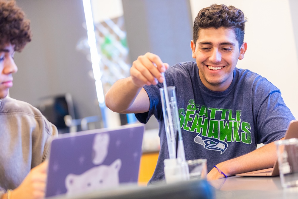 A student wearing goggles, gloves and a lab coat examines a beaker.