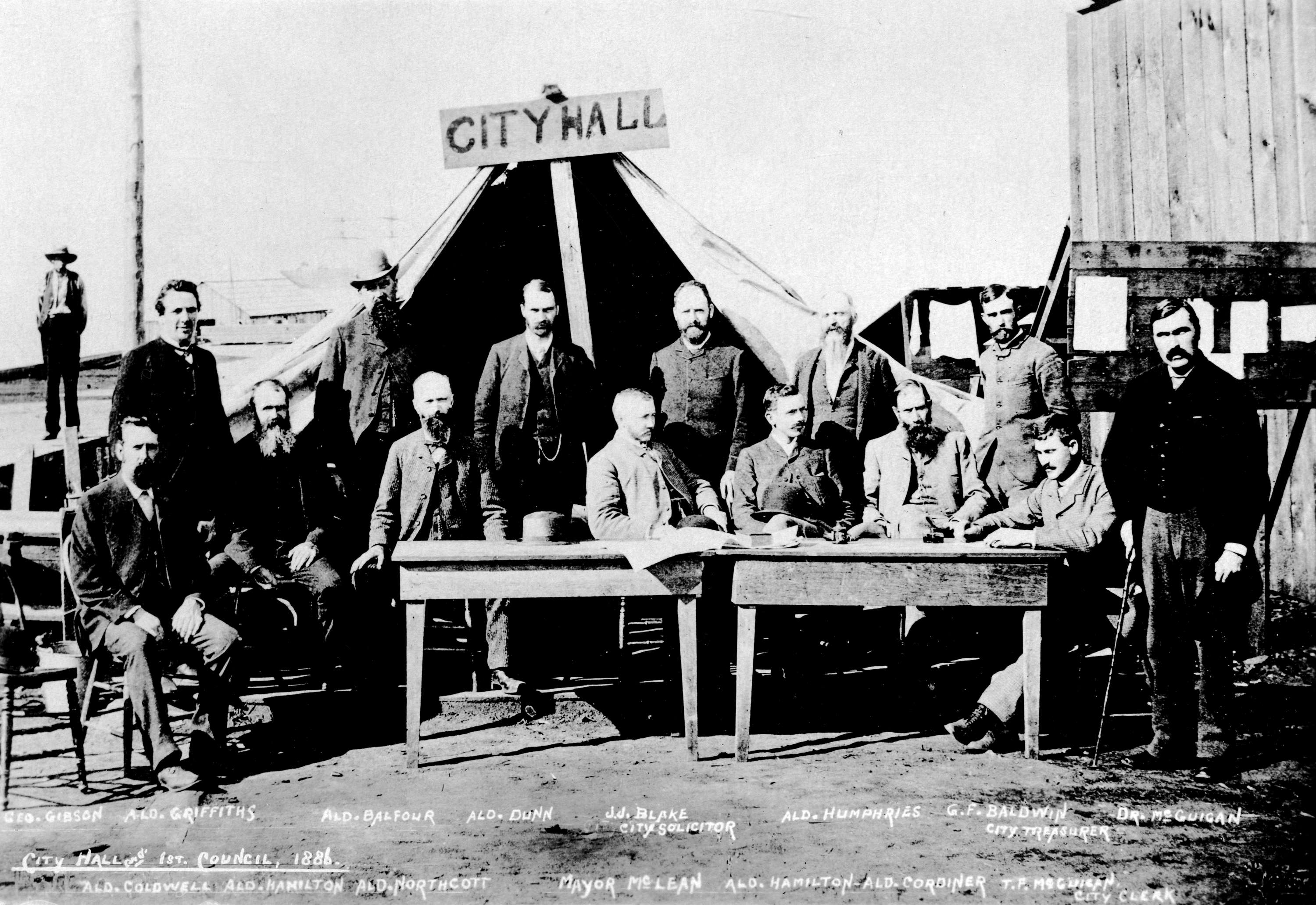 old photo of tent labeled as City Hall surrounded by about 15 men