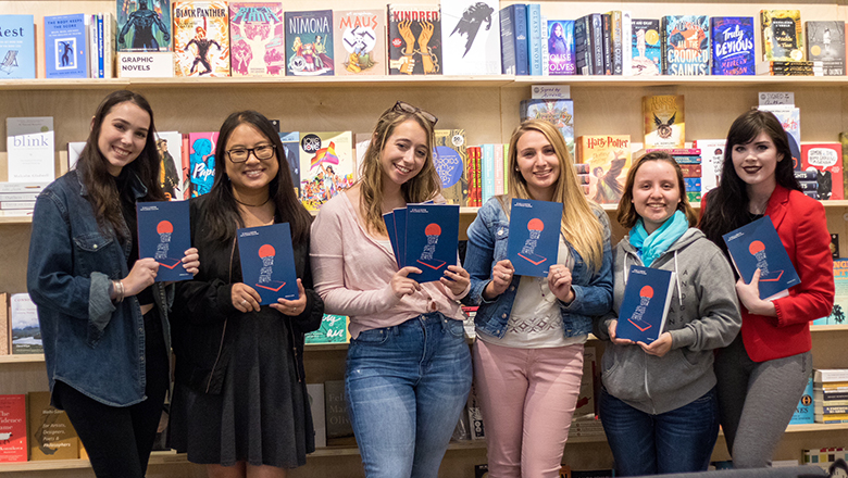 Students hold copies of a book in front of a shelf of other books in a bookstore.